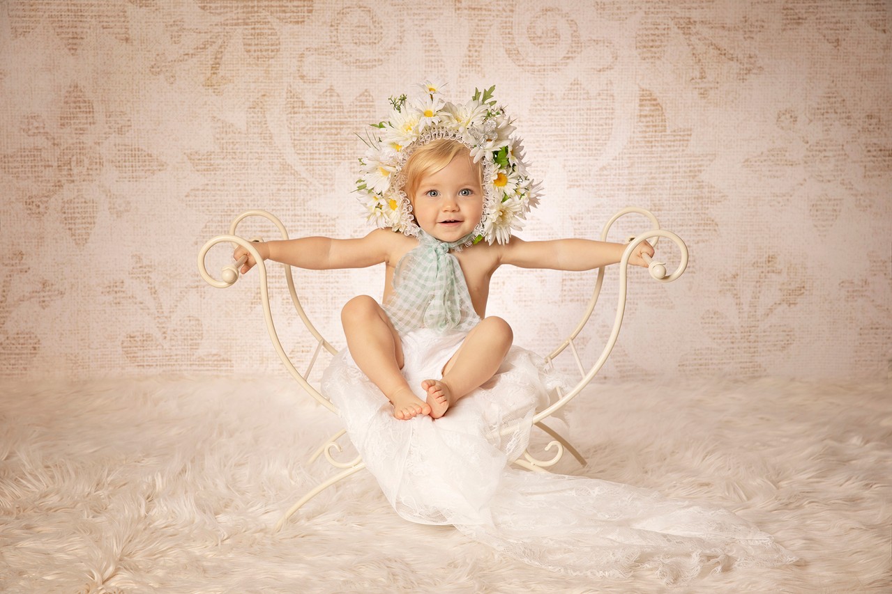 Ein Baby sitzt auf einem weißen Bettgestell und trägt einen Blumenkranz im Haar