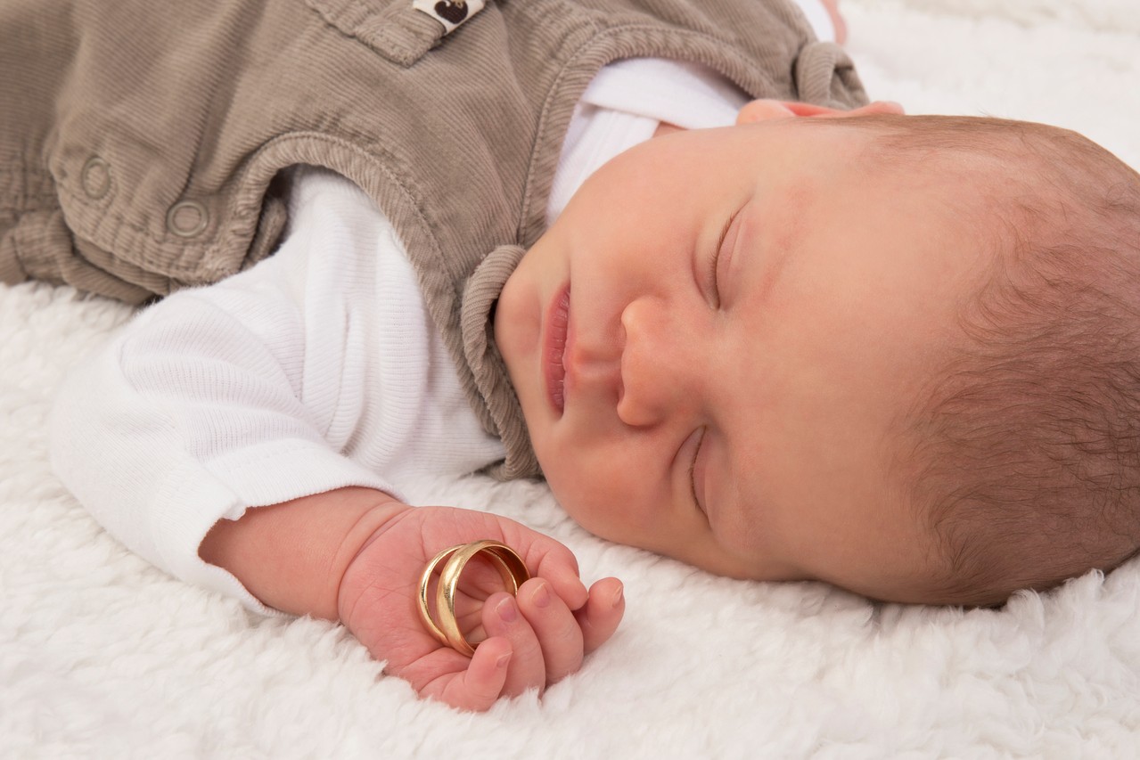 Newbornfotografie von einem schlafenden Säugling, der die Eheringe seiner Eltern in der Hand hält